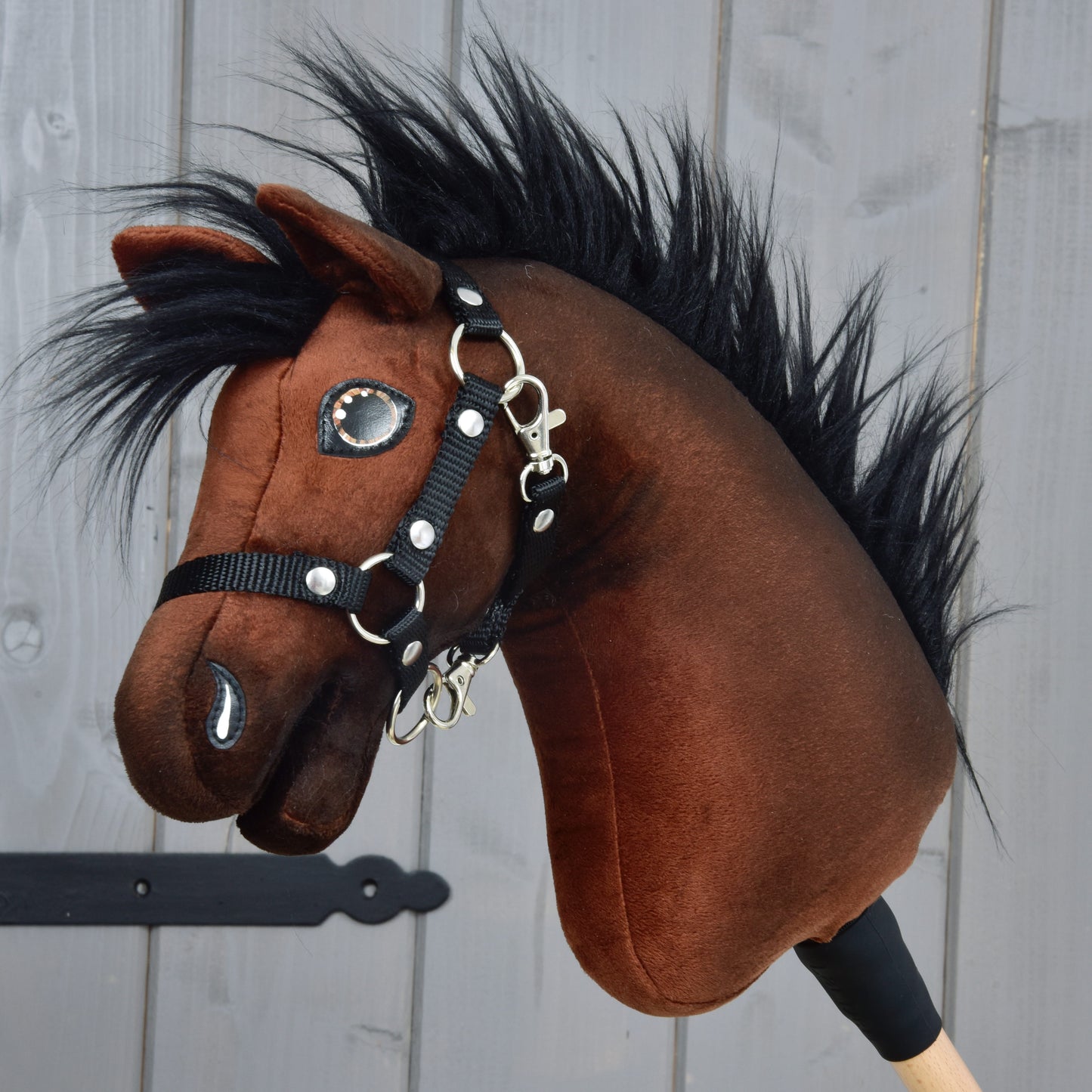 The Hobby Horse LarDen Benny with Black Halter (size XS) features a plush brown Belgian Warmblood head with black mane, white accents, a black halter, and is mounted on a wooden stick—perfect for young show jumping fans.