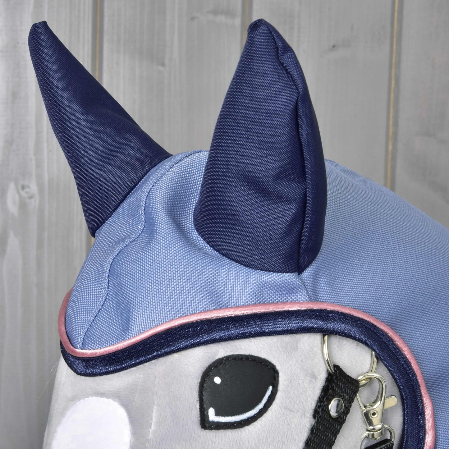 Close-up of the Hobby Horse LarDen "Raincoat Bloom," a plush horse toy sporting a light blue and navy ear bonnet with large navy ears and detailed stitching. Ideal for displaying hobby horse accessories on a wooden background.