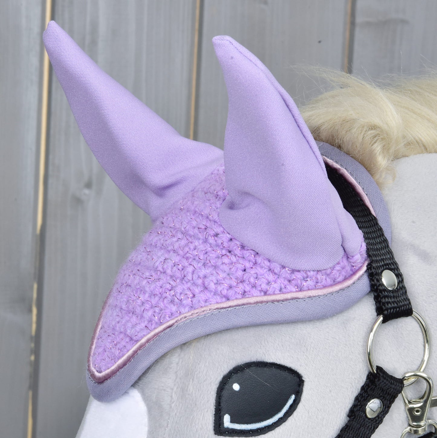 A close-up of a gray plush horse toy by Hobby Horse LarDen wearing the Orchid Ear Bonnet for insect protection and a black bridle, set against a wooden background.