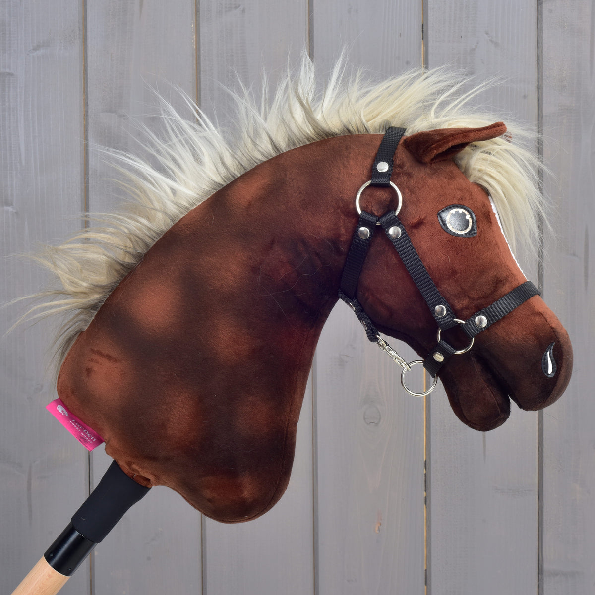 The Hobby Horse Cinnamon with Black Halter - size S by Hobby Horse LarDen, a brown plush hobby horse mounted on a wooden stick and featuring a white mane, black halter, and reins, is pictured against a gray wooden plank background.