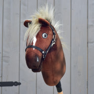 Brązowy pluszowy „Hobby Horse Cinnamon z czarnym kantarem - rozmiar S” od Hobby Horse LarDen, z białą strzałką, czarnym kantarem i puszystą blond grzywą, osadzony na drewnianym kiju na tle szarego drewnianego ogrodzenia.