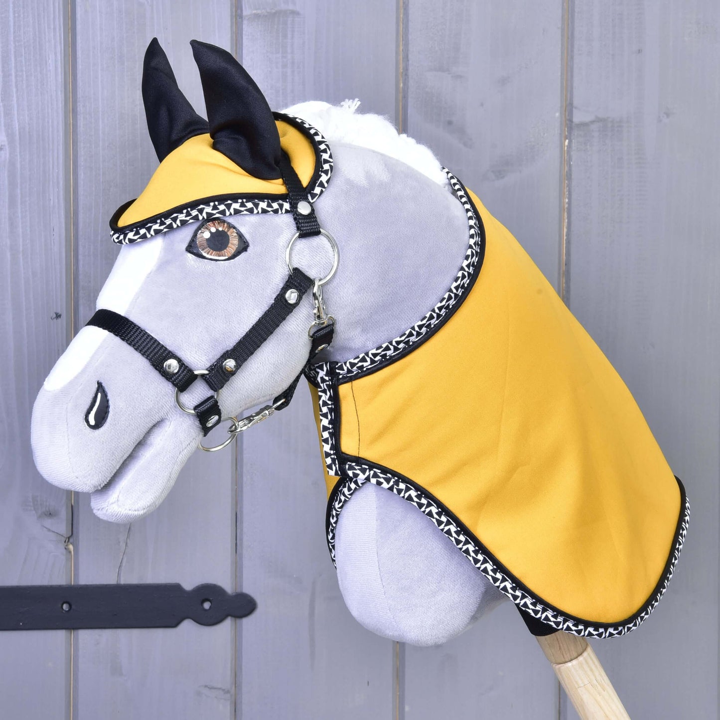 Zestaw Żółta Piękność Hobby Horse LarDen przedstawia pluszowego Hobby Horse z szarą głową, czarną uzdą, żółtym materiałowym okryciem z czarno-białym wykończeniem i pasującym czaprakiem, pokazany na drewnianym tle.