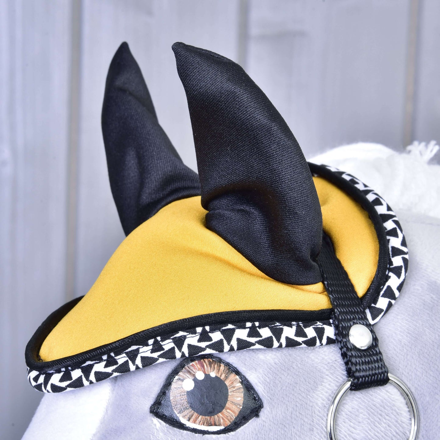 Zbliżenie na Zestaw Żółta Piękność Hobby Horse LarDen ukazuje pluszową głowę z żółto-czarnymi nausznikami, geometrycznym czarno-białym wykończeniem, malowanym okiem i białym futrzanym pyskiem na tle szarego drewna.