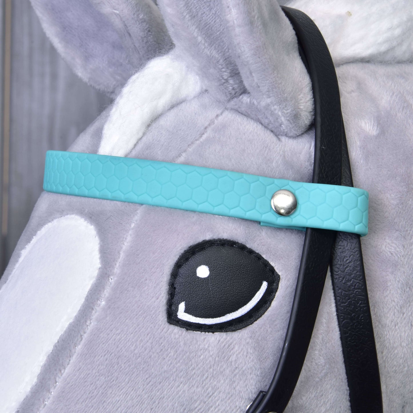 Zbliżenie głowy szarego pluszowego konia Hobby Horse LarDen noszącego turkusowe Kompletne ogłowie bez nachrapnika. Widoczne są czarna wodza, białe ucho, białe znaczenia na pysku oraz oko w stylu kreskówkowym z czarnym konturem i białym reflekssem.