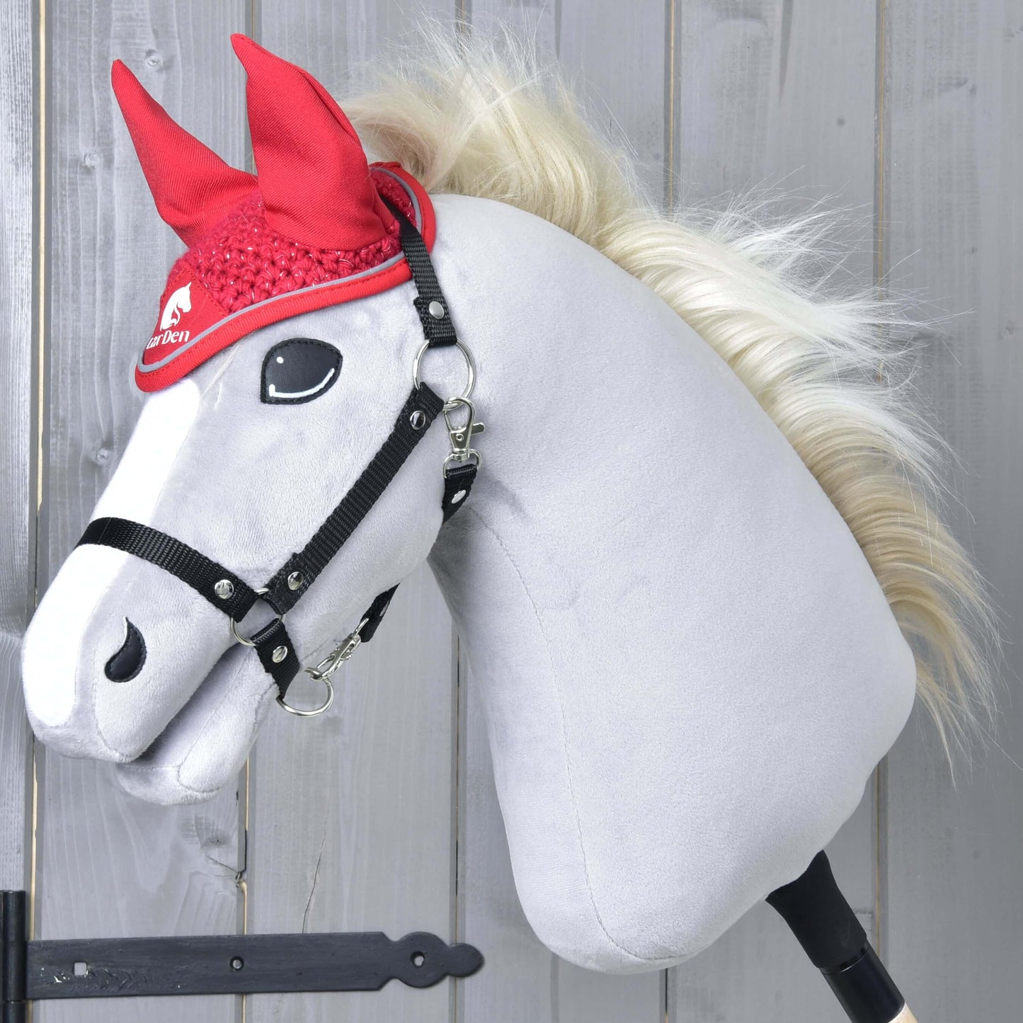 A plush hobby horse from Hobby Horse LarDen, with a white face, black bridle, and blond mane, models the Ear bonnet crocheted Red Sport in front of a light wooden wall.