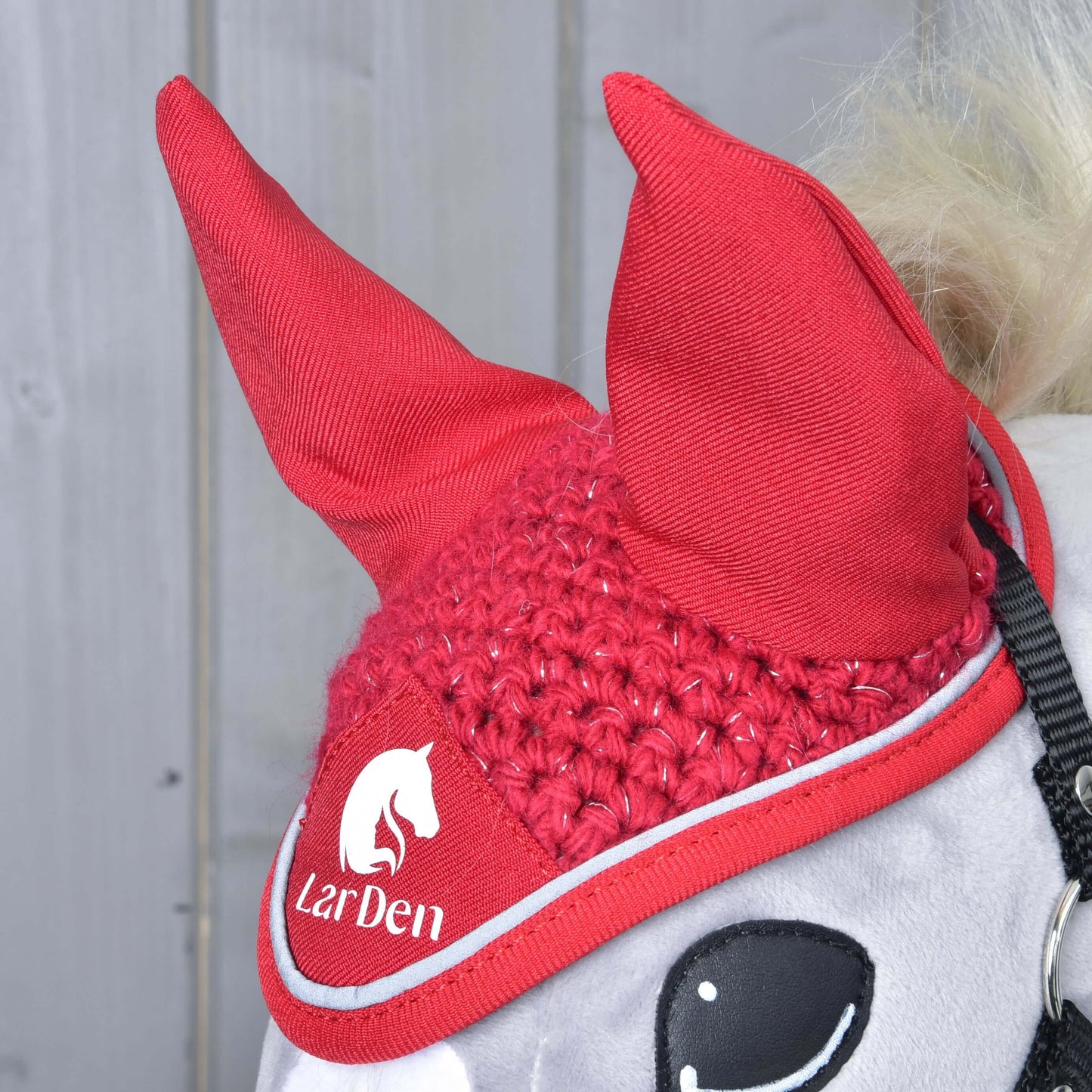 A close-up of a horse wearing the Hobby Horse LarDen Ear Bonnet Crocheted Red Sport with the white LarDen logo and horse graphic, set against light gray wooden panels.
