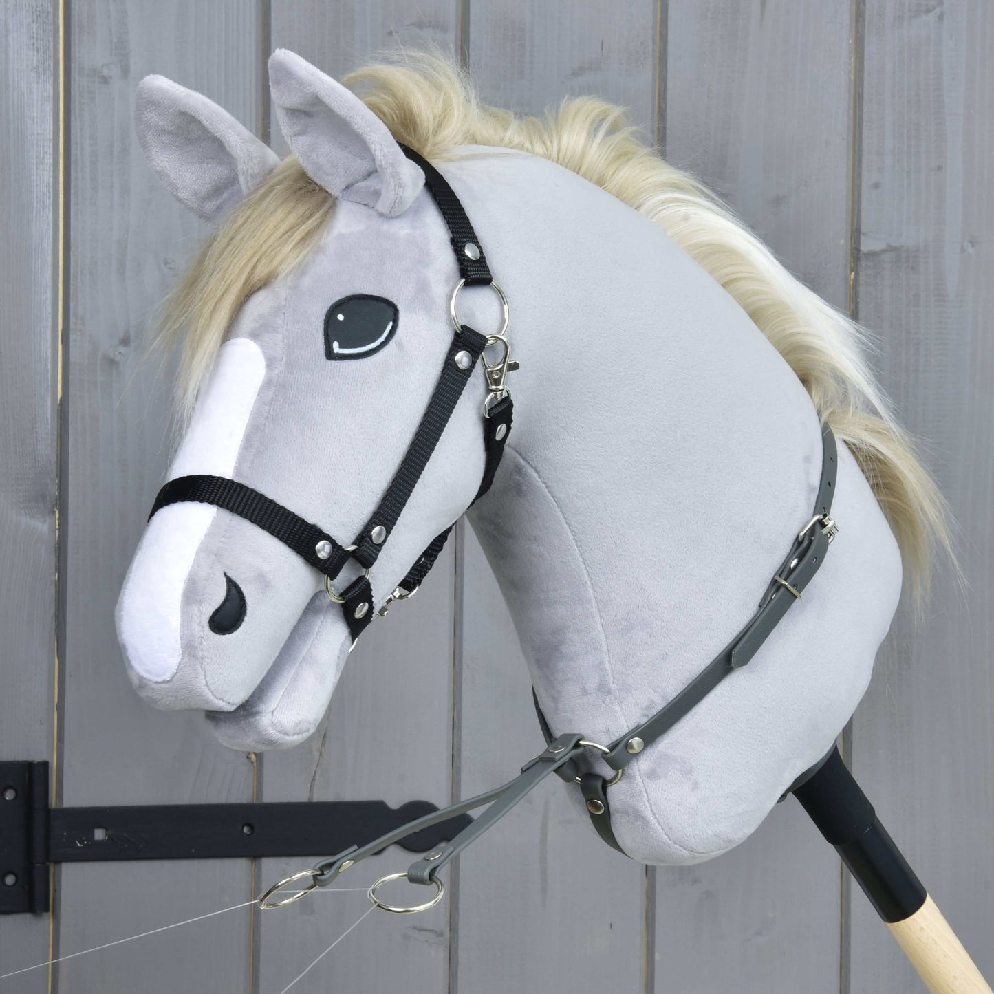A close-up of the Hobby Horse LarDen "Martingale dark grey" plush hobby horse with a white mane, black bridle, and leatherette martingale, mounted on a wooden stick in front of a gray wooden wall.