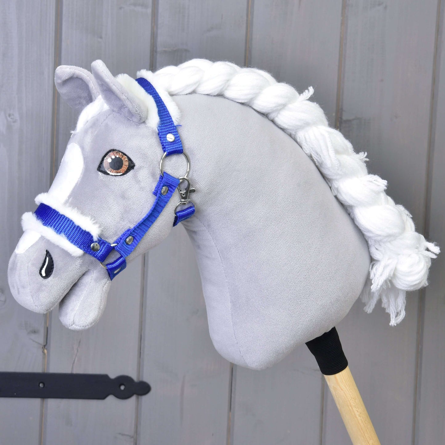 A gray fabric hobby horse with a white yarn mane and the "Hobby Horse LarDen" Halter blue with fur stands in front of a light gray wooden wall.