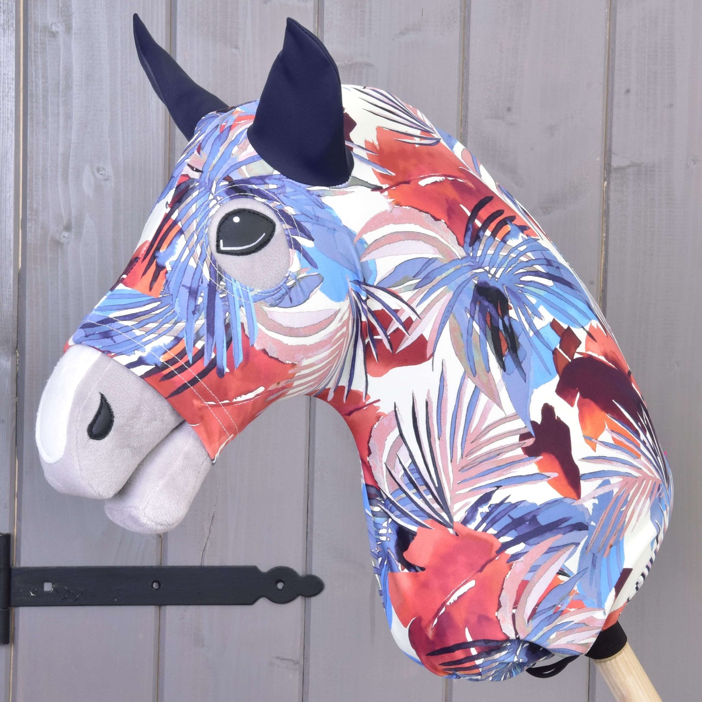 The Hobby Horse LarDen Hobby Horse Protective Suit Blue Jungle (Lycra) features a vibrant red, white, purple, and blue abstract pattern with black eyes and dark ears. Includes a washable cover and is shown on a wooden stick by a gray wall.