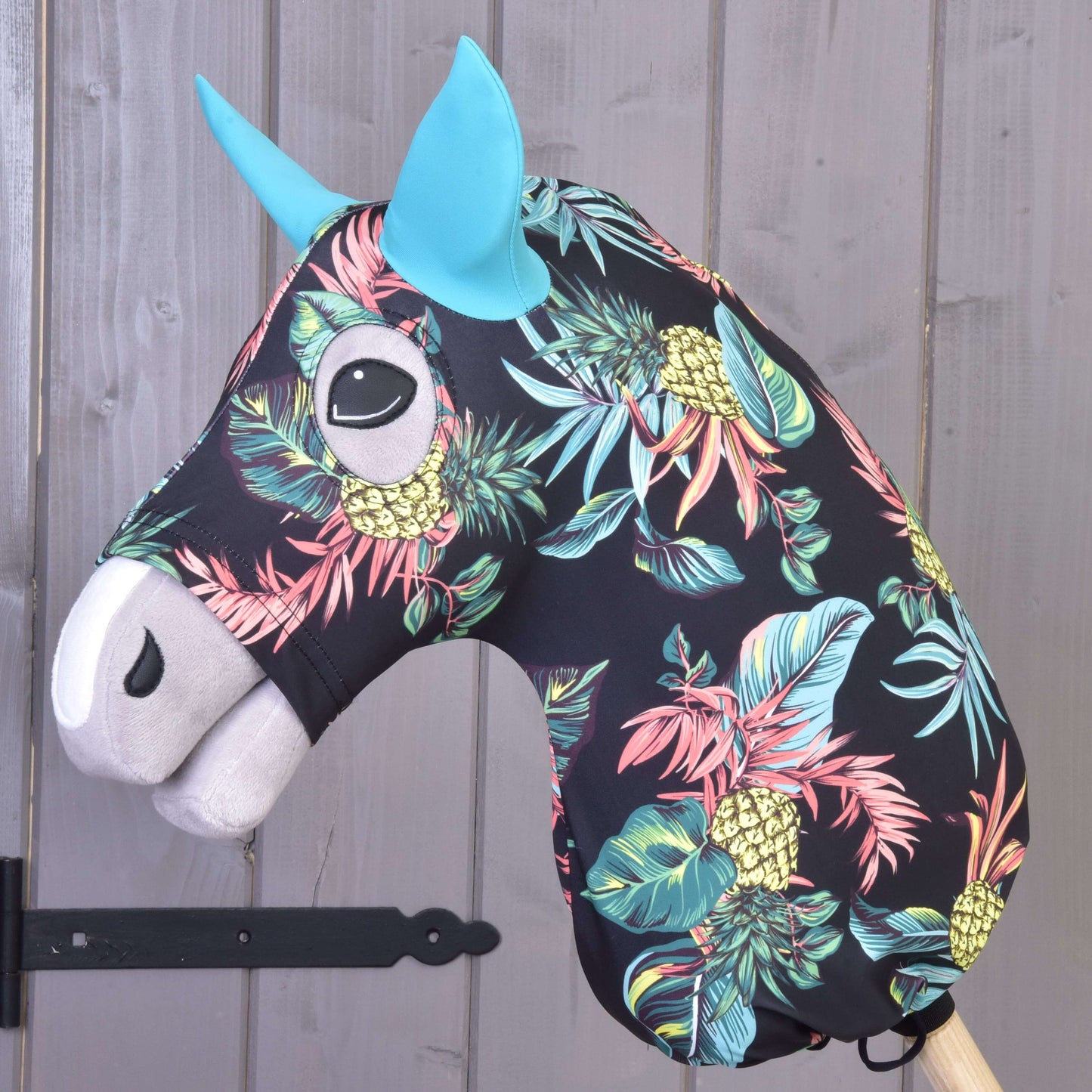 The Hobby Horse LarDen’s Protective suit Pineapple (Lycra) features a black head with a vibrant pineapple and leaf print, blue ears, and a white muzzle, mounted on a wooden stick and displayed against a gray wooden wall.