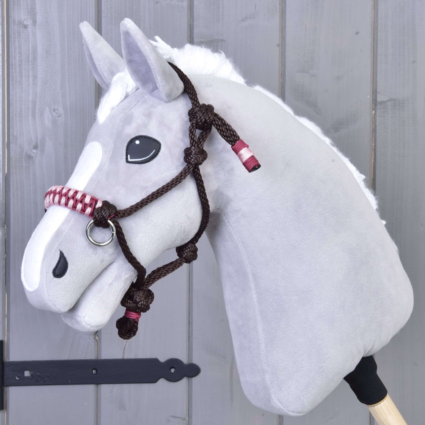 The Hobby Horse LarDen Rope halter York, a gray plush hobby horse head with a white mane and nose patch plus a brown braided rope halter, hangs on a gray wooden wall.