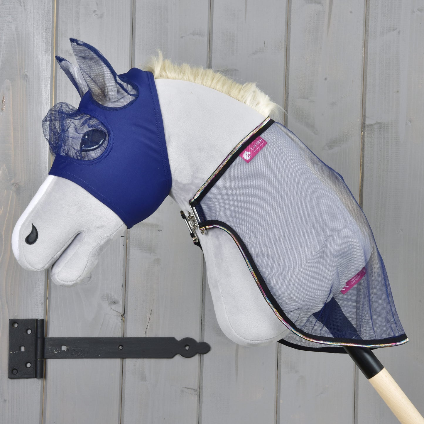 Hobby Horse LarDen Zestaw Fly Combo przedstawia pluszowego Hobby Horse z niebieską maską przeciw owadom i siatkową derką przeciw owadom na drewnianym kiju, na tle szarej drewnianej ściany, gotowego na kolejną przygodę.