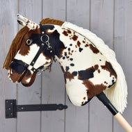 Hobby Horse Dakota z czarnym kantarem od Hobby Horse LarDen, z brązowymi, czarnymi i białymi łatami, brązową grzywą z włóczki i czarnym kantarem, osadzony na drewnianym kiju na tle szarej ściany; wzór odpowiedni dla każdej płci.