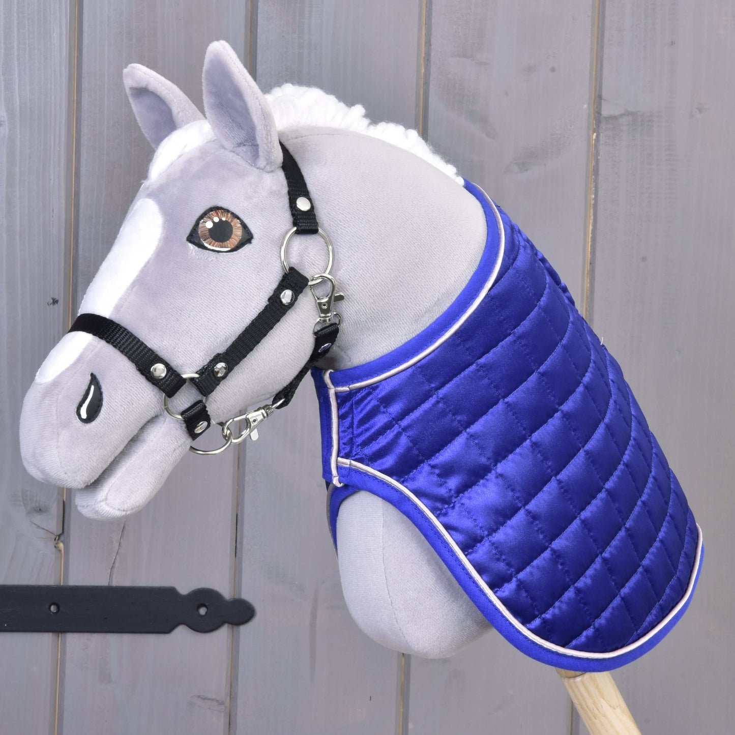 Pluszowy Hobby Horse z białą grzywą, czarnym kantarem i derką Hobby Horse LarDen Derka DeLuxe niebieska dwustronna dla dodatkowej ochrony jest osadzony na drewnianym kiju na tle szarego drewna.