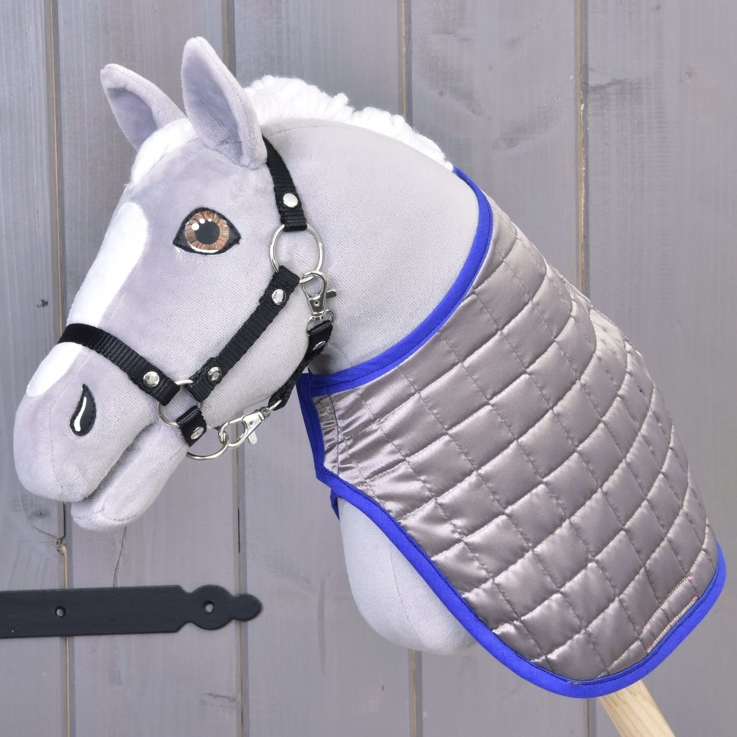 Szary pluszowy Hobby Horse z czarną uzdą i dwustronną derką Derka DeLuxe niebieska od Hobby Horse LarDen, na tle szarego drewna.