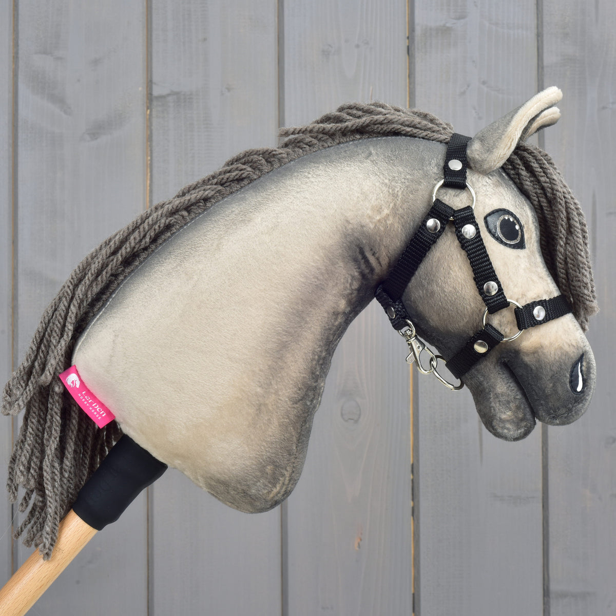 The Hobby Horse LarDen "Hobby Horse Dusty with Black Halter" (size XS) features a realistic gray plush Hanoverian head, detailed with button eyes and a black halter, and is mounted on a wooden stick. It is shown positioned against a wooden plank background.