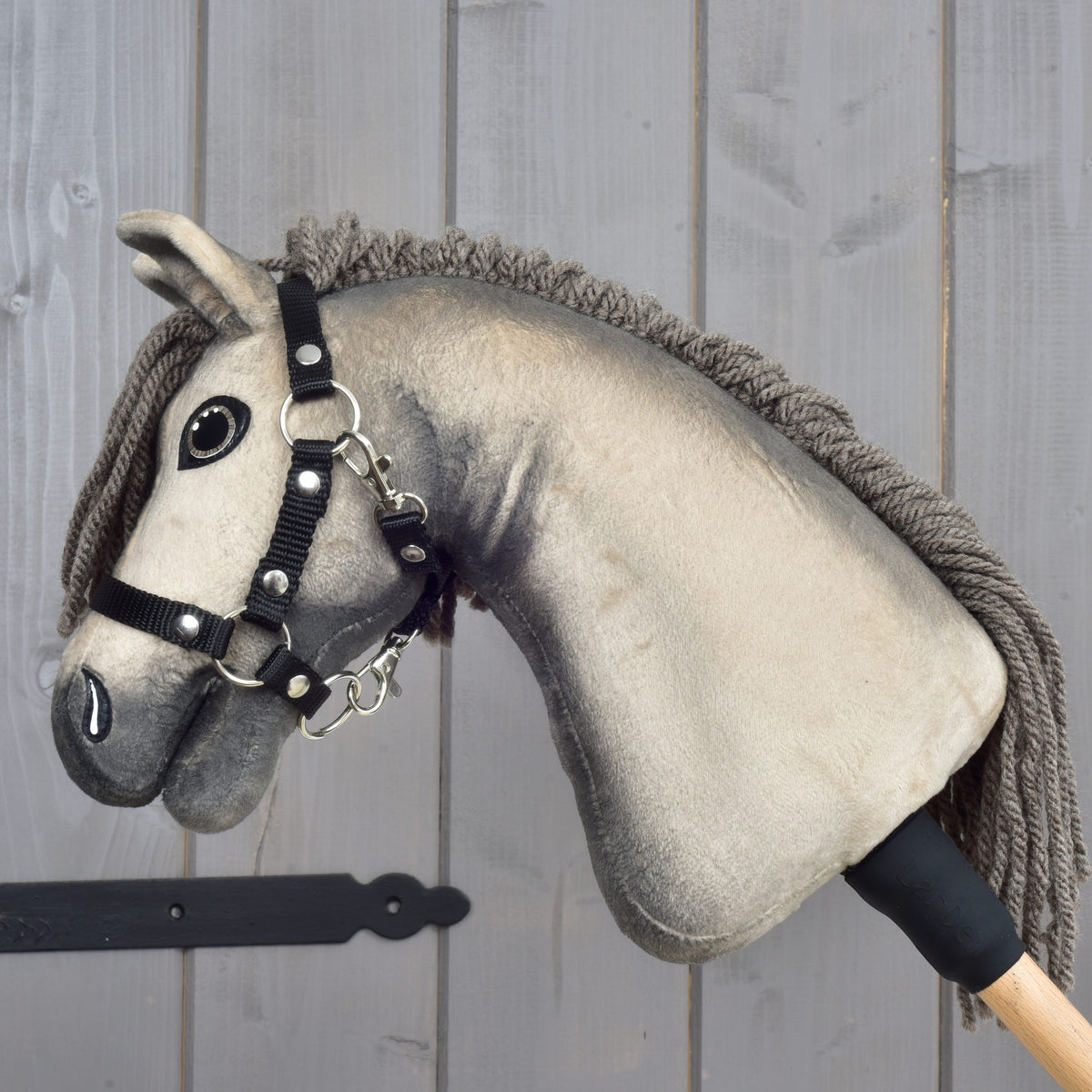 The Hobby Horse LarDen "Hobby Horse Dusty with Black Halter" (size XS) features a plush Hanoverian head with button eyes and a black halter, mounted on a wooden stick and set against a light wooden panel background.