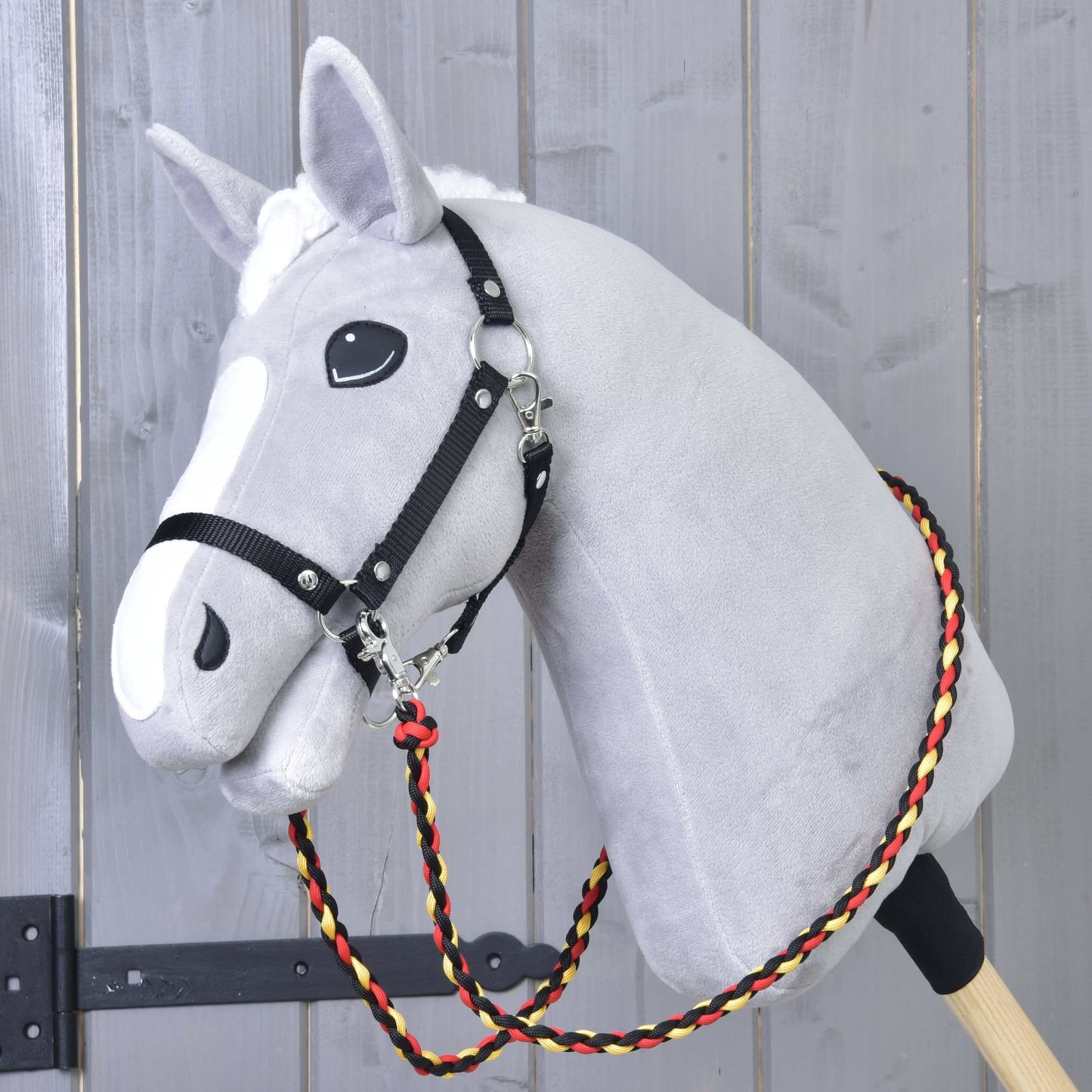 Hobby Horse LarDen Reins Flag to szary pluszowy Hobby Horse na drewnianym kiju, z czarną uprzężą i wodzami handmade z plecionego czerwonego, żółtego i czarnego sznurka polipropylenowego, pokazany na tle drewnianej ściany.