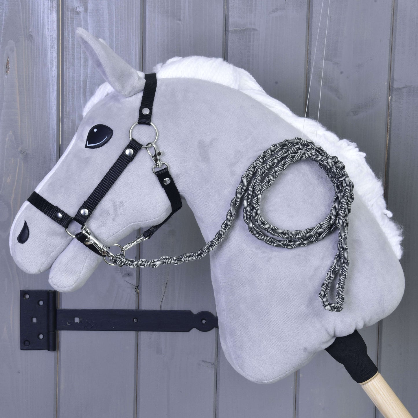 Hobby Horse - Dziana lonża Graphite od Hobby Horse LarDen przedstawia szarą pluszową głowę, czarną uzdę i ręcznie dzianą lonżę Graphite na drewnianym kiju, pokazane na tle szarej drewnianej ściany.