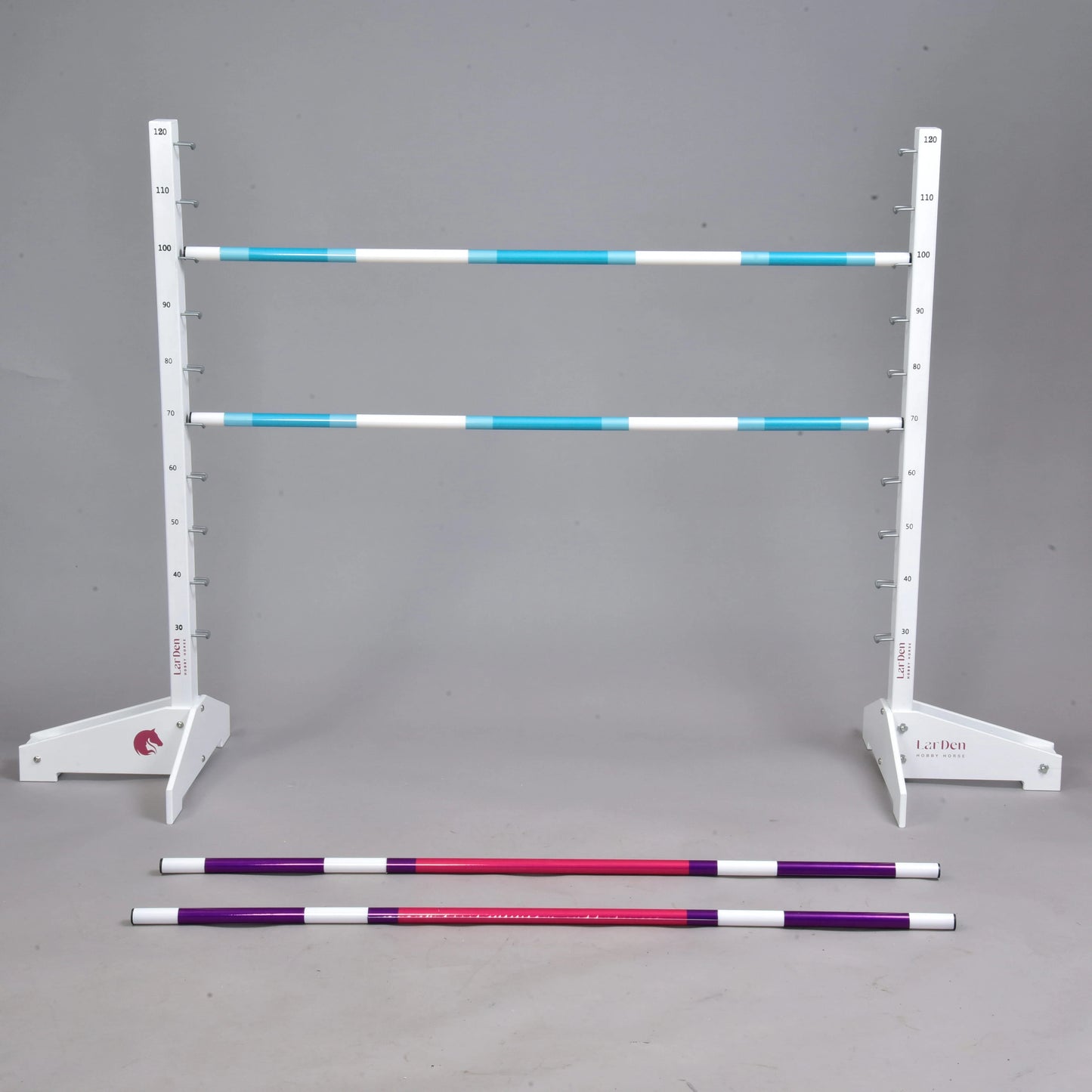 Przeszkoda 120 cm / 47 inch z dodatkowymi tyczkami od Hobby Horse LarDen, z białym regulowanym stojakiem podtrzymującym dwie niebiesko-białe tyczki w paski oraz czerwone i fioletowe tyczki na szarej podłodze. Idealna do Hobby Horsing, na jednolitym szarym tle.