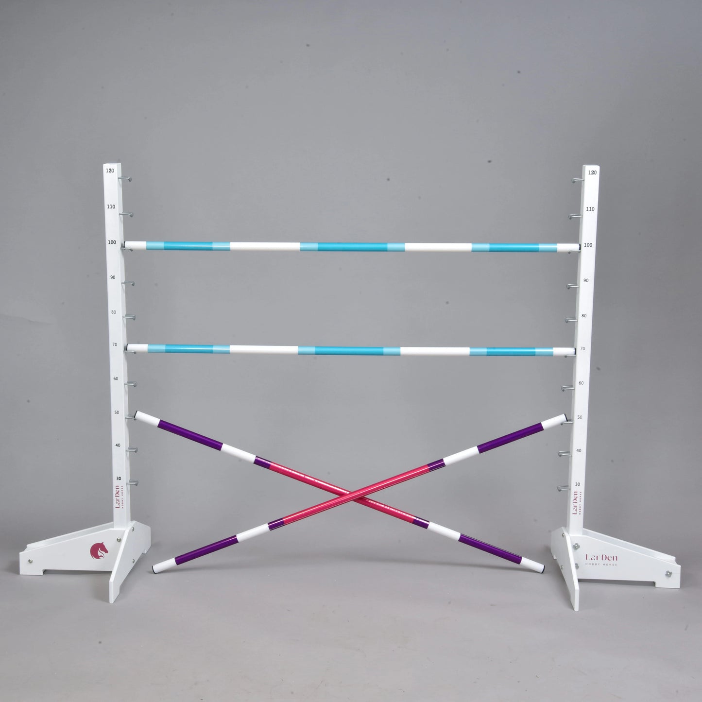 Przeszkoda 120 cm / 47 inch z dodatkowymi tyczkami od Hobby Horse LarDen, zawiera dwa białe stojaki, trzy regulowane niebiesko-białe tyczki oraz dwie skrzyżowane fioletowo-białe tyczki i jest idealna do Hobby Horsing w pomieszczeniach lub na zewnątrz. Pokazana na jednolitym szarym tle.
