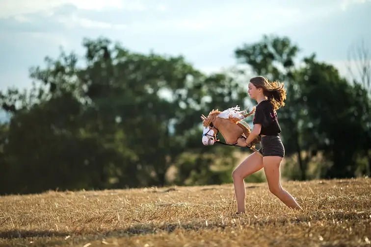 A young person runs through a dry, grassy field while hobby horsing with a toy hobby horse, with trees and a blue sky in the background.