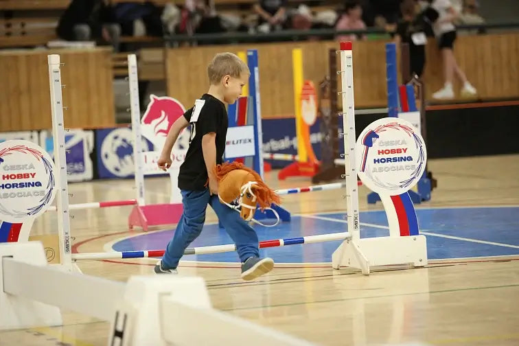 A young boy takes part in a Hobby Horse competition, walking with a toy horse on a gym floor, surrounded by colorful jumping obstacles and hobby horsing event signs.