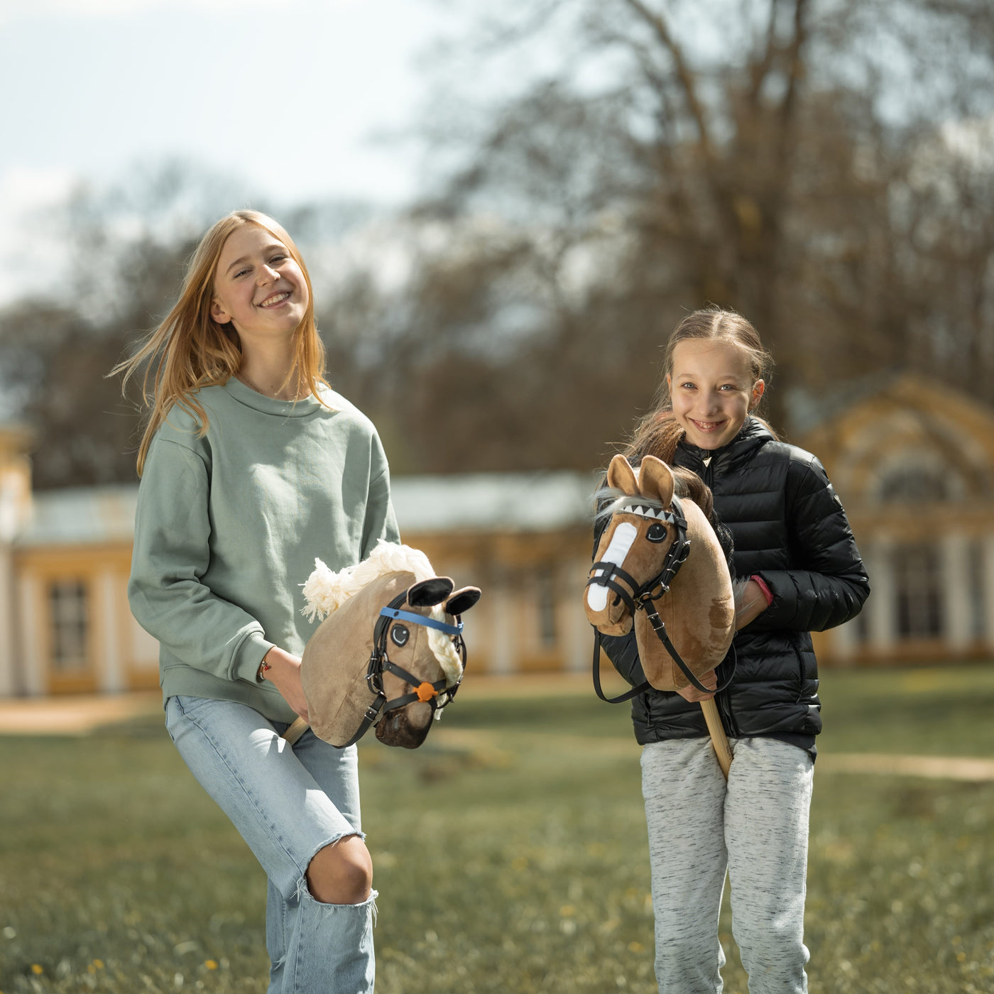 Dwie uśmiechnięte dziewczynki stoją na zewnątrz, trzymając swoje hobby horse. Jedna ma na sobie jasnzielony sweter i podarte dżinsy, druga czarną kurtkę i szare spodnie. Hobby Horsing sprawia im radość, gdy pozują w pobliżu dużego budynku i drzew w tle.