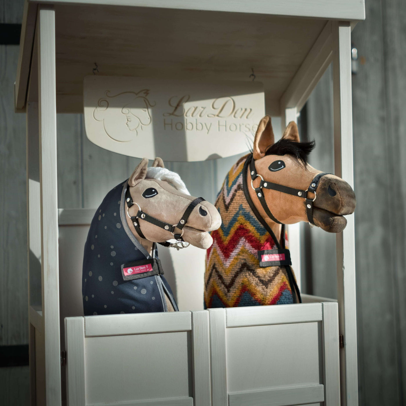 Dwa pluszowe Hobby Horse z uzdami i derkami, jeden w niebieskim pokrowcu w kropki, a drugi w kolorowy wzór zygzaków, stoją obok siebie w białej drewnianej budce z napisem LarsDen Hobby Horsing.