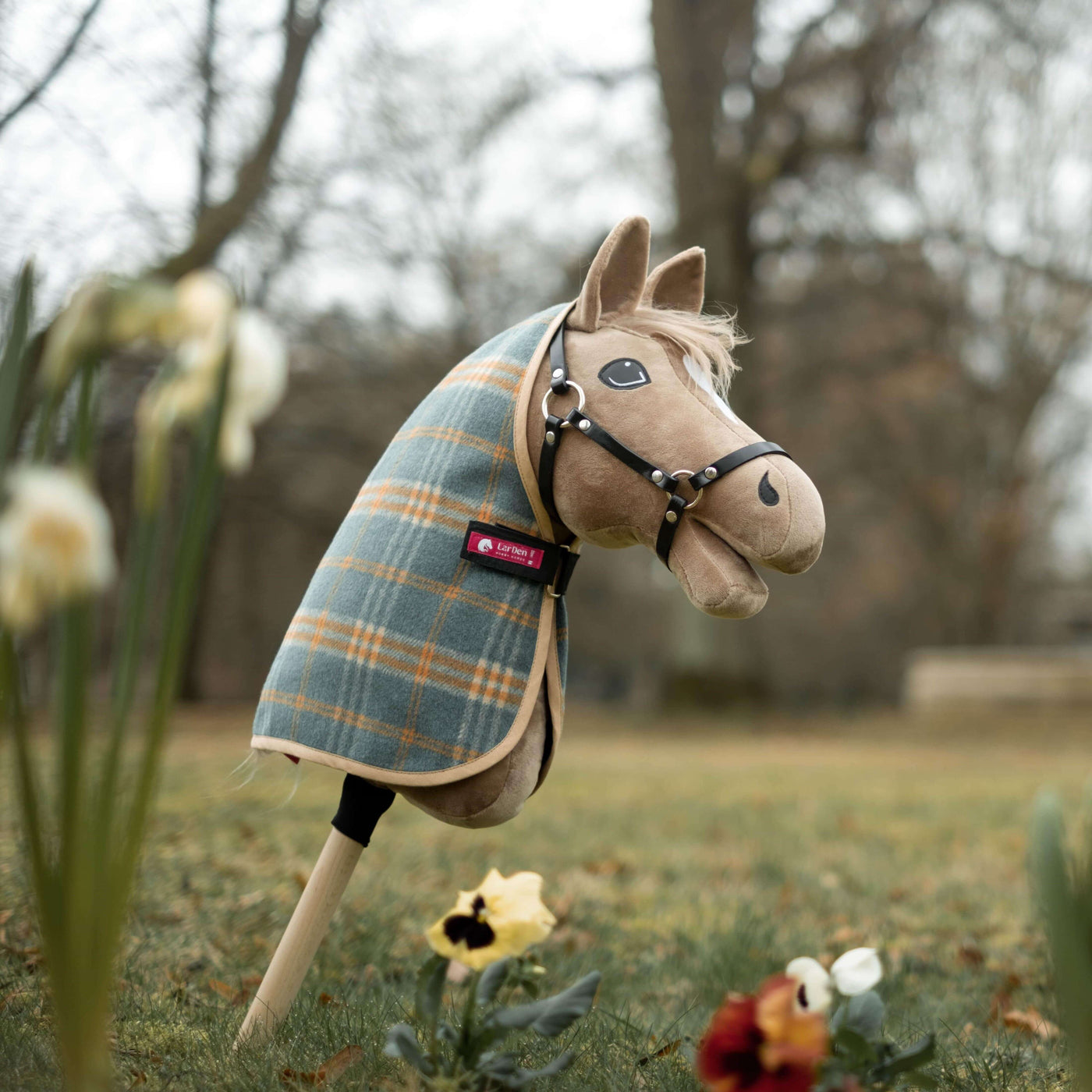 Pluszowy Hobby Horse w derce w kratę i uzdzie stoi pionowo na kiju na trawiastym terenie na zewnątrz, idealny do przygód z Hobby Horsing wśród wiosennych kwiatów i drzew w tle.