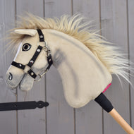 A Hobby Horse LarDen “Hobby Horse Ivory Dream with Black Halter” in size XS, featuring soft ivory fur and a wooden stick handle, is leaning against a gray wooden wall.