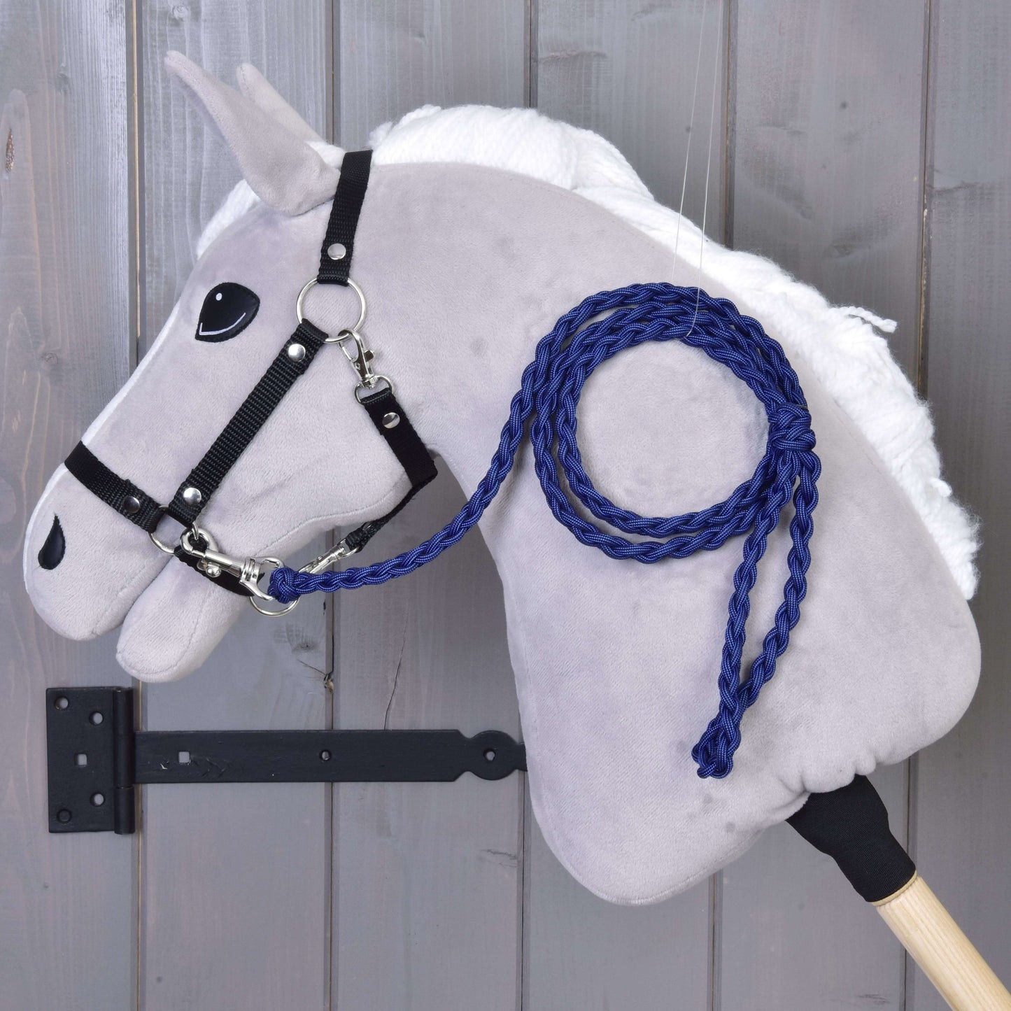 A gray plush Hobby Horse - Knitted Lead Rope Royal Blue by Hobby Horse LarDen, featuring a white mane, black bridle, and royal blue hand-knitted lead rope, is mounted on a wooden wall with its stick secured by a black metal bracket.