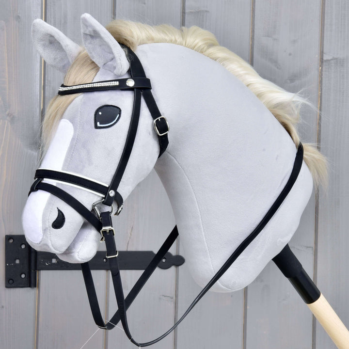Kompletne ogłowie De Luxe z 2 naczółkami Hobby Horse LarDen na realistycznej szarej zabawce Hobby Horse na kiju z białą grzywą i nachrapnikiem kombinowanym, osadzonej na drewnianym kijku na tle szarego drewna.