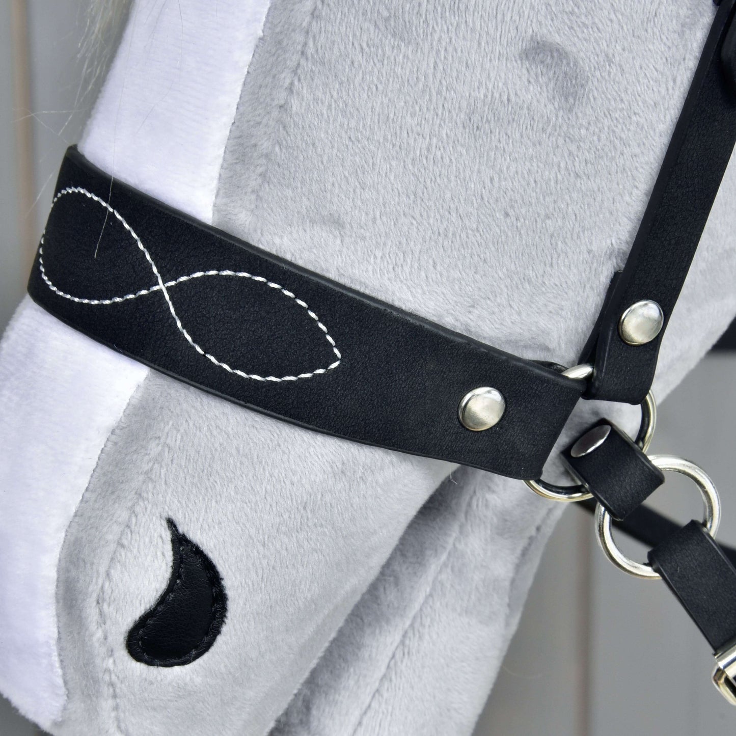 Close-up of a grey plush hobby horse’s head wearing the Hobby Horse LarDen Complete bitless bridle, decorated with white infinity-symbol stitching and a teardrop-shaped black patch near its eye.