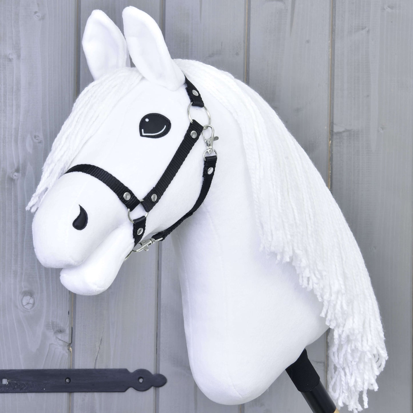 Hobby Horse Lady z czarnym kantarem od Hobby Horse LarDen, przedstawia białego konia z czarnym kantarem i długą włóczkową grzywą na kiju, upozowanego jak gotowy do ujeżdżenia na tle jasnoszarej drewnianej ściany.
