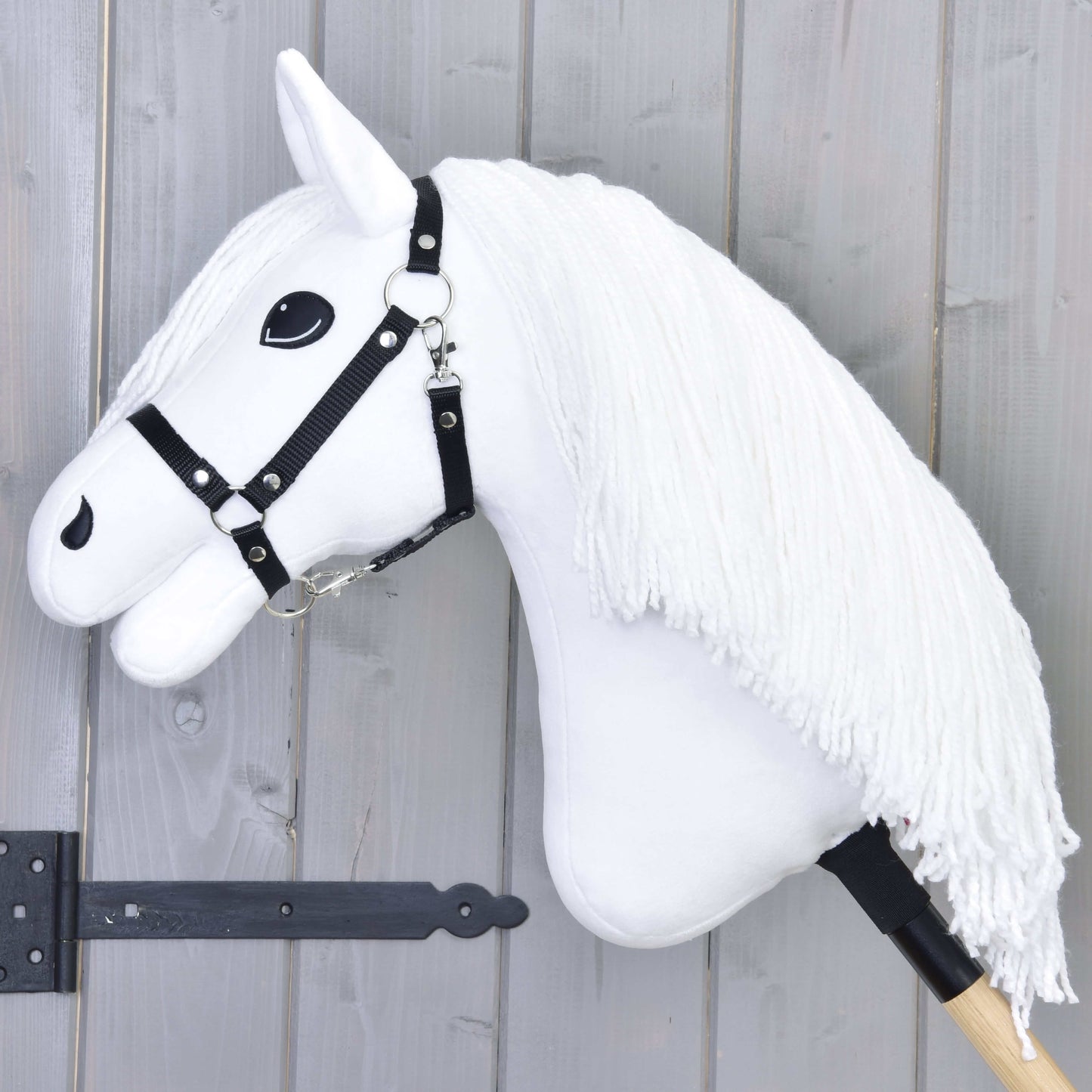 Hobby Horse Lady z czarnym kantarem od Hobby Horse LarDen z białą końską głową, długą włóczkową grzywą, czarnym kantarem i wyrazistymi czarnymi oczami na drewnianym kiju, pokazany na tle szarego drewna.