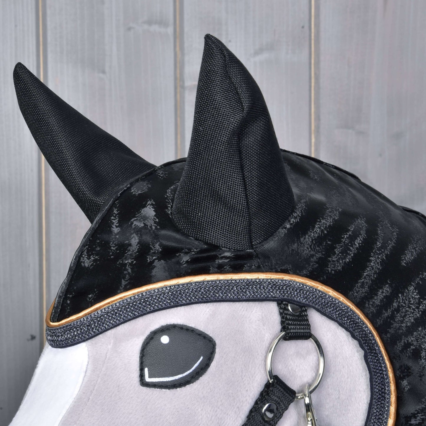 Close-up of the Hobby Horse LarDen Raincoat Luxury: a black and gray fabric hobby horse head with large black ears, stitched smiling eye, shiny black mane, and a metal ring by the mouth, set against wooden panels.