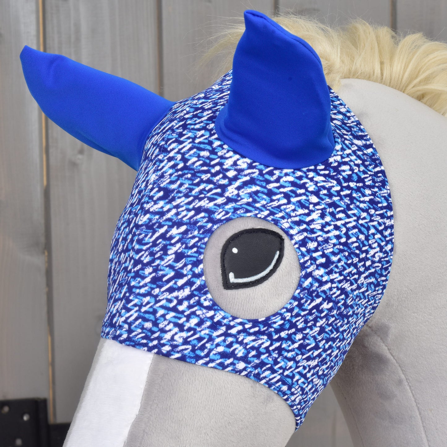A close-up of a plush horse’s head wearing the Hobby Horse LarDen Horse Mask See, featuring blue ears and a blue and white pattern, set against a wooden background.