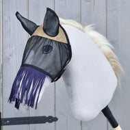 The Hobby Horse LarDen Fly Mask De Luxe Mona is shown on a white plush hobby horse with a black stick handle and light blonde mane, featuring a stylish fringe fly mask against a gray wooden background.