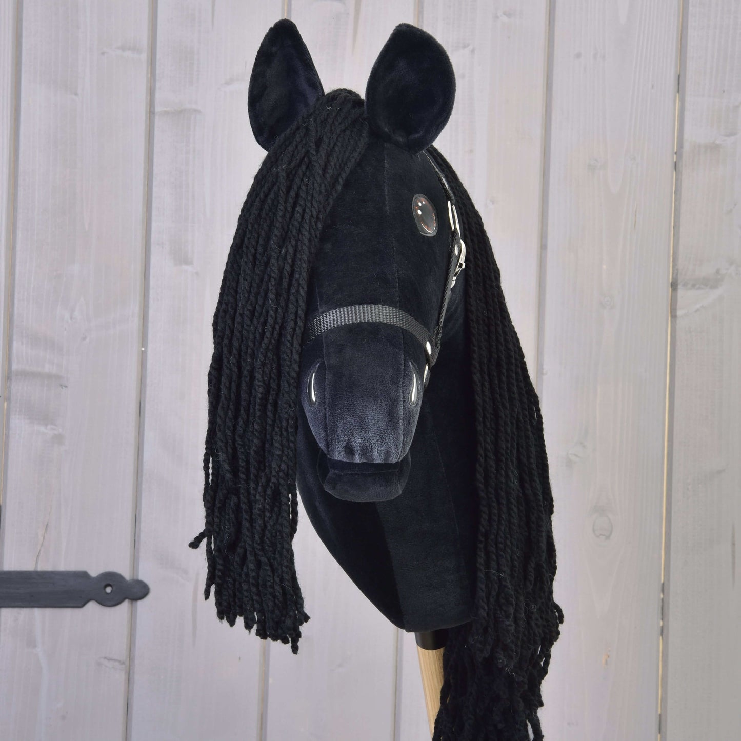 Hobby Horse LarDen’s Maximus with Black Halter is a handmade Friesian hobby horse featuring a plush head, yarn mane, button eyes, black halter, and wooden stick, shown here against a light wood background.