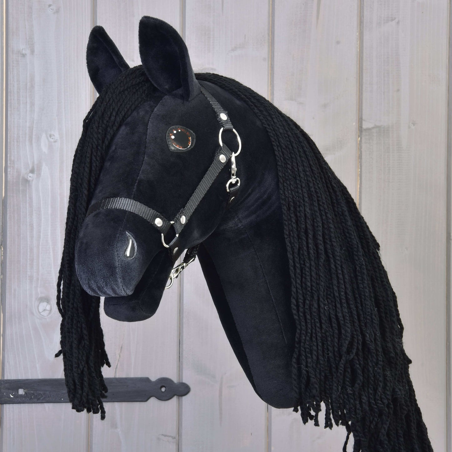 The Hobby Horse LarDen Maximus with a black halter is a plush black Friesian hobby horse with a long black mane, mounted on a wooden stick and shown against a light wood-paneled background.