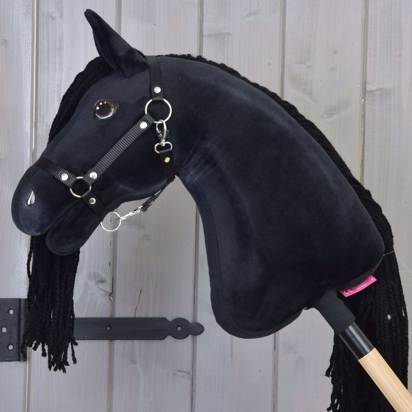 The Hobby Horse LarDen's Hobby Horse Maximus with Black Halter features shiny button eyes and a yarn mane, mounted on a wooden stick, displayed in front of a light wooden plank wall.