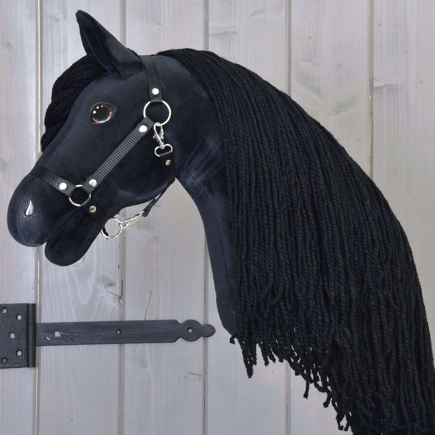 The Hobby Horse Maximus with Black Halter by Hobby Horse LarDen, featuring a plush black Friesian design, long braided mane, bridle with metal rings, and shiny button eye, hangs on a wooden wall.