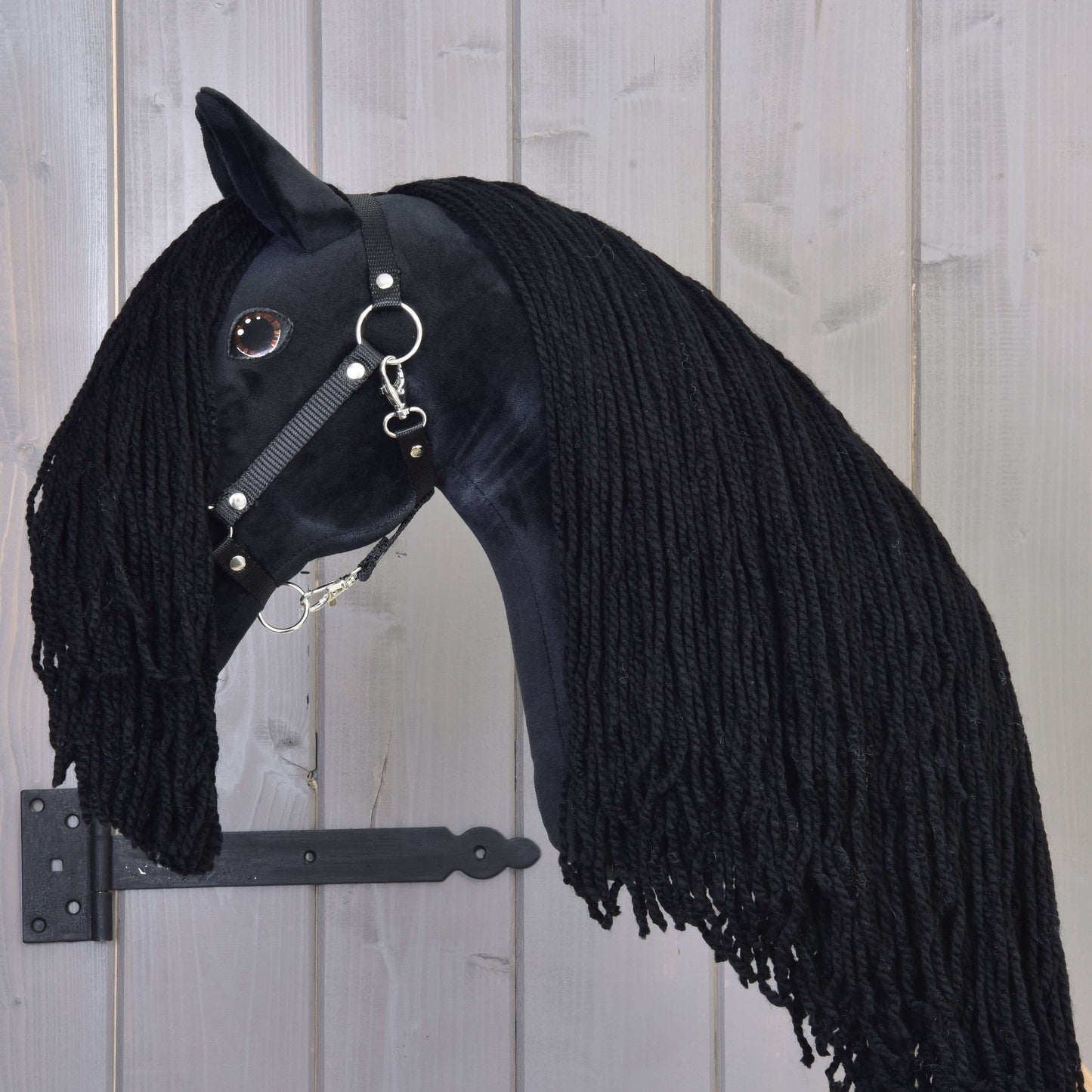 The Hobby Horse Maximus with Black Halter by Hobby Horse LarDen is a handmade black stallion toy featuring a long yarn mane, shiny black bridle, and button eye, mounted on wood with a black metal bracket.