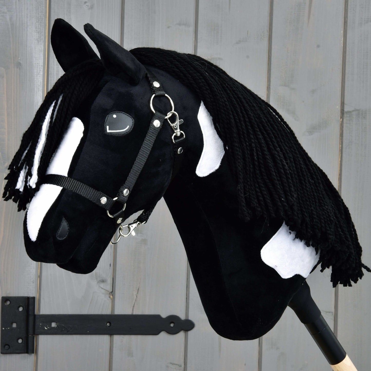 The Hobby Horse LarDen "Hobby Horse Merlin with Black Halter," inspired by a Barock Pinto stallion, features black and white spots and a braided yarn mane, mounted against a gray wooden wall.