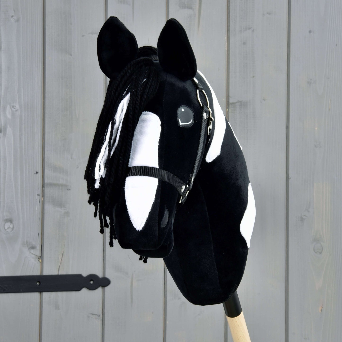 The Hobby Horse Merlin with Black Halter by Hobby Horse LarDen is a black-and-white spotted hobby horse styled after a Barock Pinto stallion, featuring a yarn mane and bridle, mounted on a wooden stick in front of a gray fence.