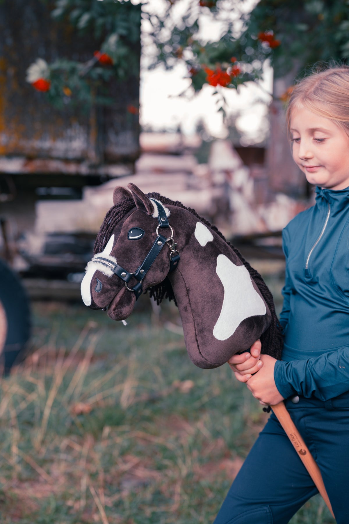 Młoda osoba w granatowym stroju jeździeckim trzyma Hobby Horse Monte od Hobby Horse LarDen na łące przed starym drewnianym wozem i jarzębiną z czerwonymi owocami, z brązowo-białą sierścią inspirowaną American Paint Horse, długą ciemną grzywą z włóczki i kantarem Espresso z futrzanym podszyciem