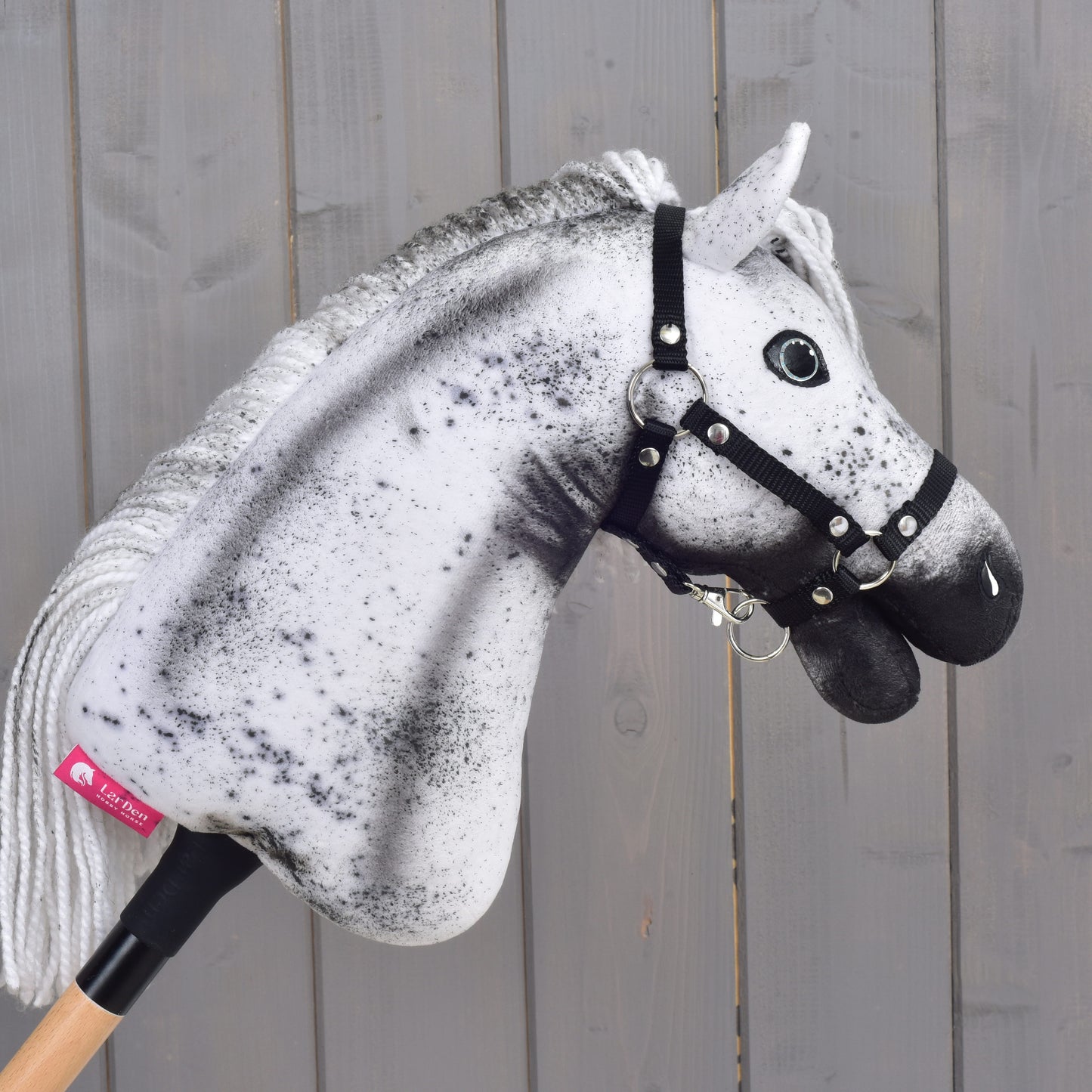 Hobby Horse Naivy z czarnym kantarem - rozmiar S od Hobby Horse LarDen, z wyrazistym biało-szarym nakrapianym wzorem, osadzony na drewnianym kiju na tle szarej drewnianej ściany.