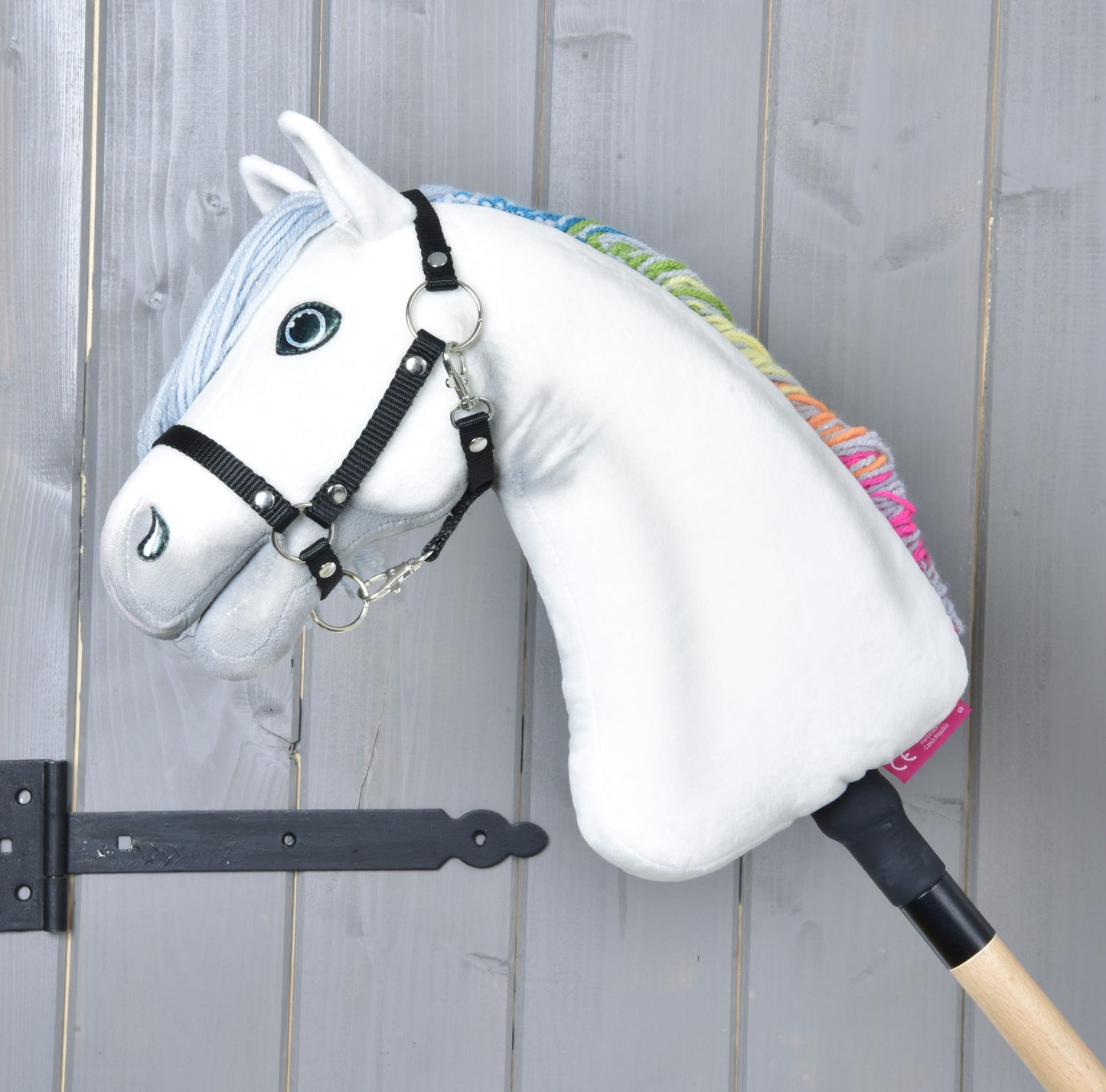 The Hobby Horse Rainbow New by Hobby Horse LarDen features a white toy horse with a black bridle and rainbow mane on a wooden stick, shown leaning against a gray wooden wall.
