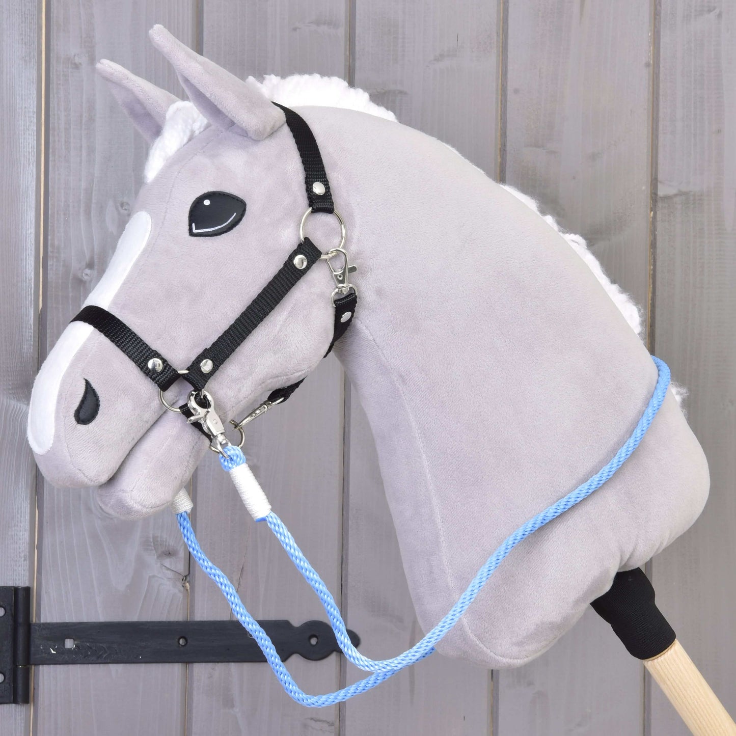 Wodze Sky Hobby Horse LarDen: szary pluszowy Hobby Horse z czarną uzdą, niebieską liną polipropylenową i ręcznie robionymi wodzami na drewnianym kiju, pokazany przed drewnianą ścianą.